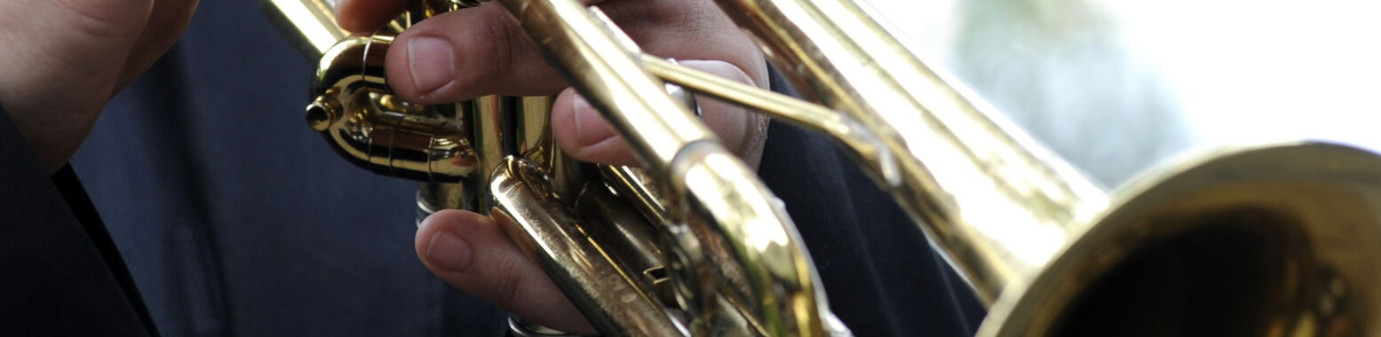 Primo piano delle mani di un musicista in abito scuro che suona una tromba dorata; le dita sono appoggiate sulle valvole.