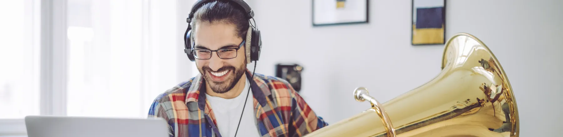 Ein lächelnder Mann mit Kopfhörern sitzt mit einer großen Tuba im Arm an einem Schreibtisch vor seinem Laptop, in einer Online-Musikstunde.