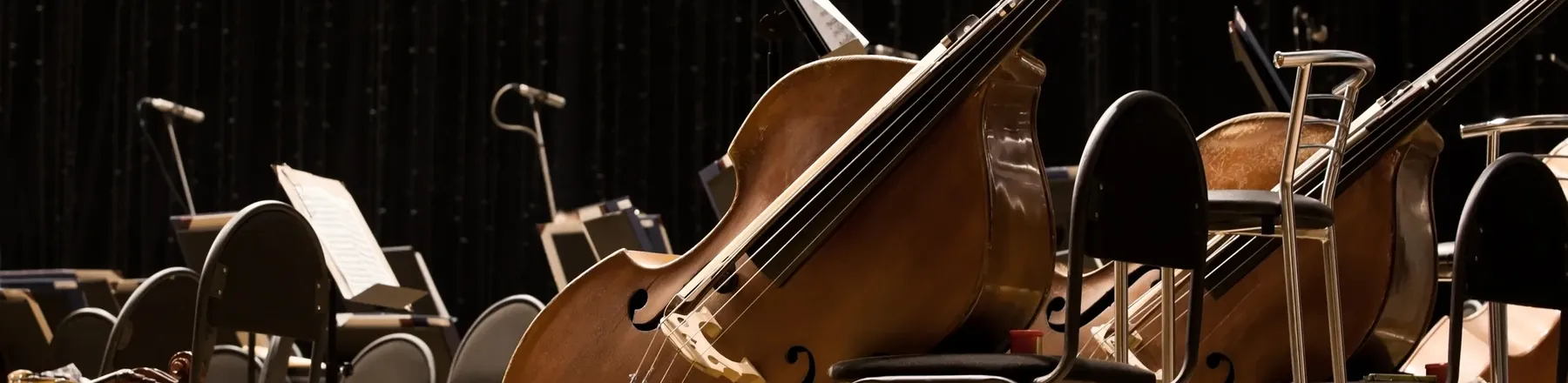 Mehrere Kontrabässe lehnen unbenutzt an Stühlen auf einer dunklen Orchesterbühne, umgeben von leeren Notenständern.