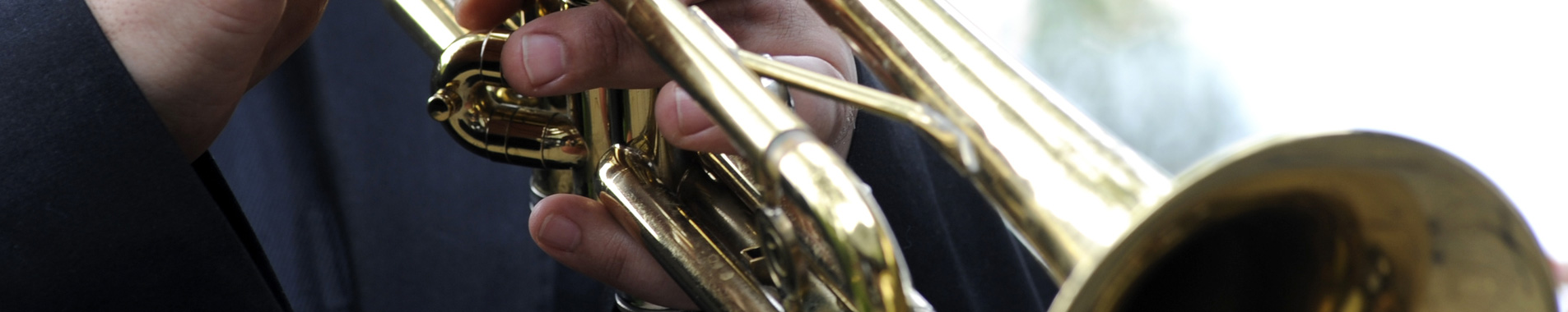 Primo piano delle mani di un musicista in abito scuro che suona una tromba dorata; le dita sono appoggiate sulle valvole.