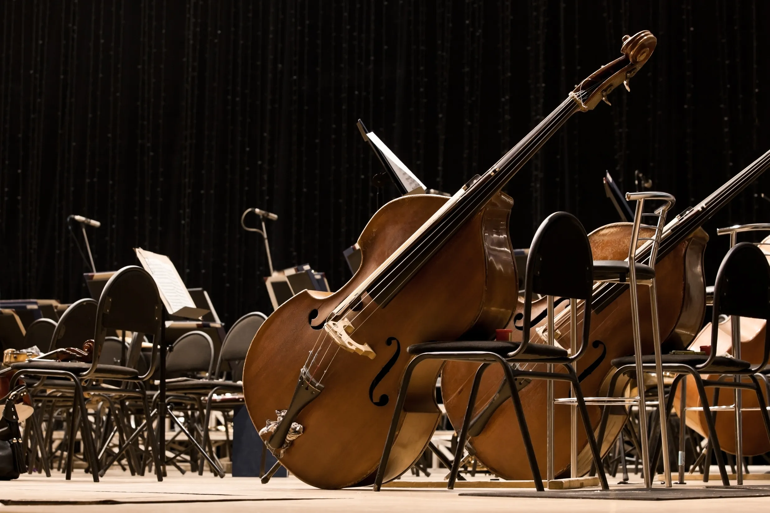 Mehrere Kontrabässe lehnen unbenutzt an Stühlen auf einer dunklen Orchesterbühne, umgeben von leeren Notenständern.