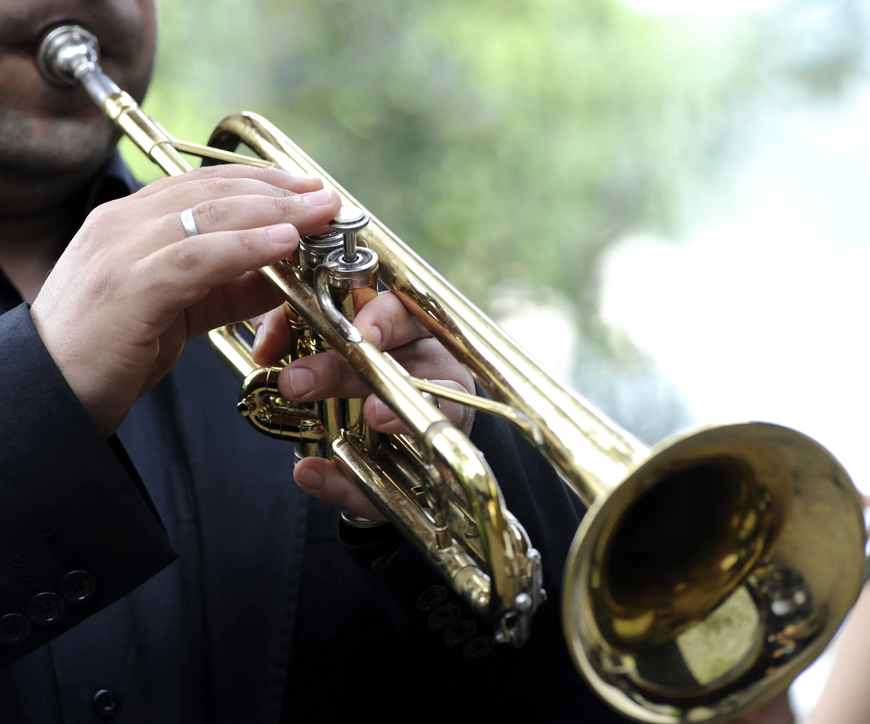 Nahaufnahme der Hände eines Musikers im dunklen Anzug, der eine goldene Trompete spielt; die Finger liegen auf den Ventilen.